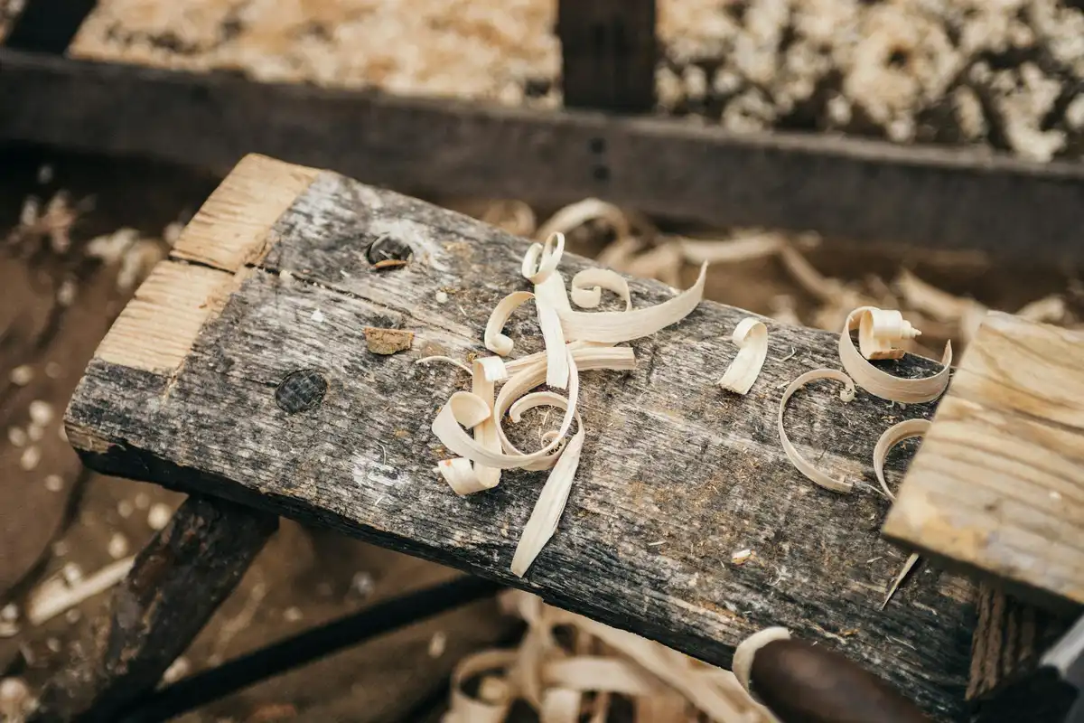 Close-up of curly wood shavings on a rough, weathered wooden bench with a few metal marks and sawdust.