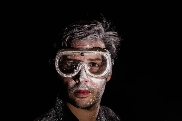 Person wearing protective goggles with white residue on their face against a dark background.