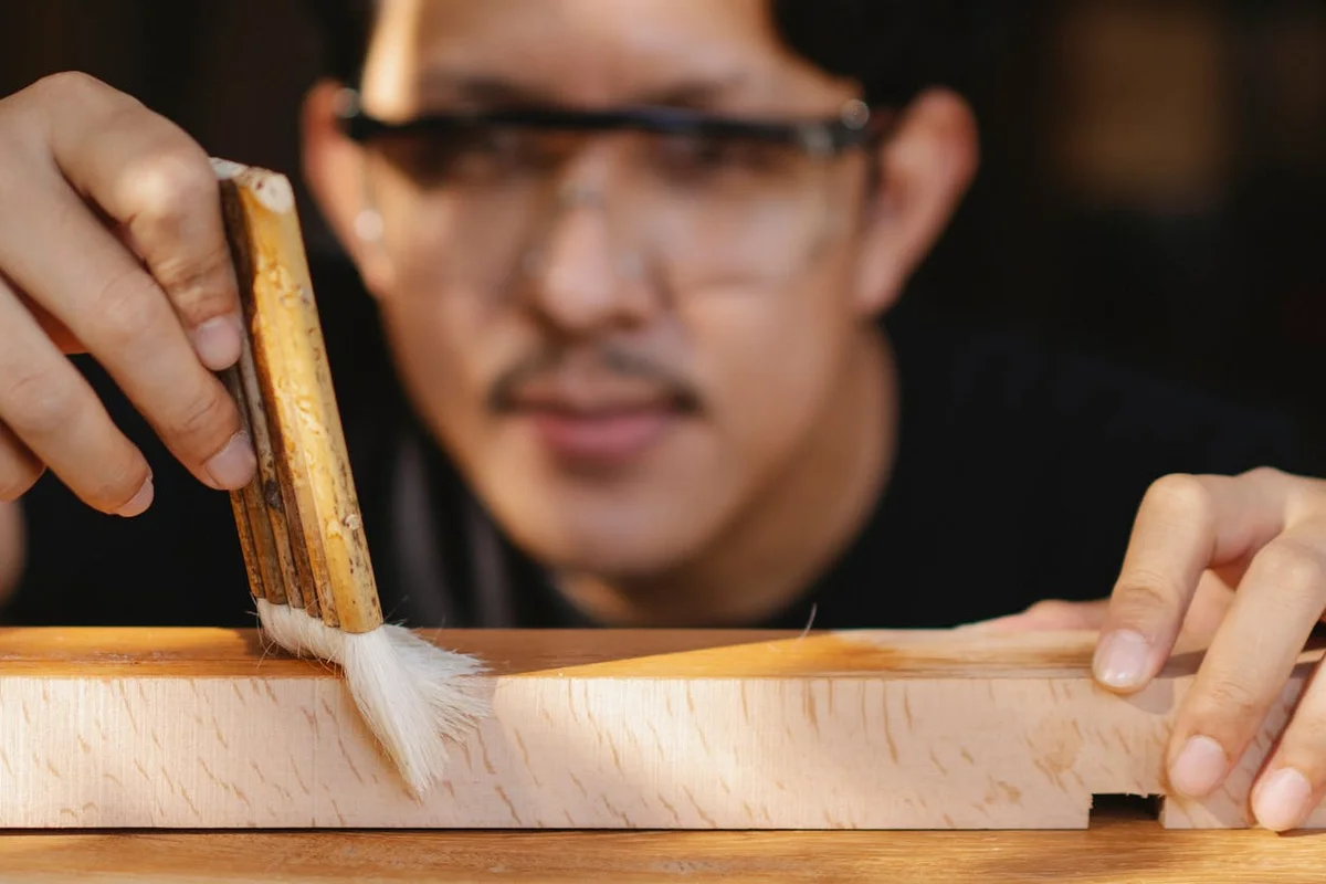 Close-up of a craftsman in safety glasses applying wood stain to a wooden plank with a brush, focusing on pigment penetrating the wood.