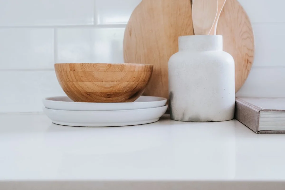 Minimal kitchen setup with a wooden salad bowl atop white plates, a white ceramic oil bottle, and cutting boards on a clean white countertop