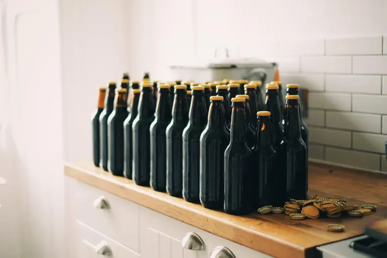 Row of dark glass bottles with cork caps lined up on a wooden kitchen countertop beside a tiled backsplash.