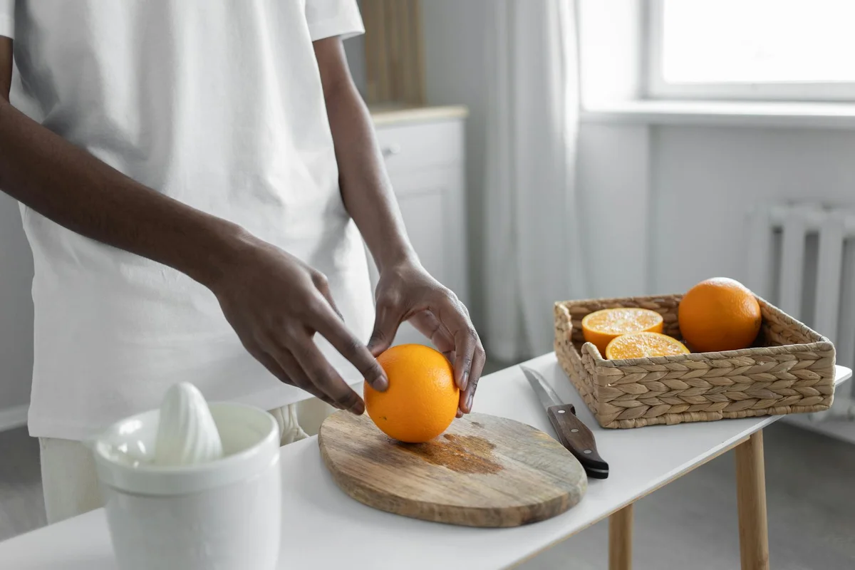 Person slicing an orange on a wooden cutting board in a bright kitchen, with a knife resting on the board and a basket of oranges in the background