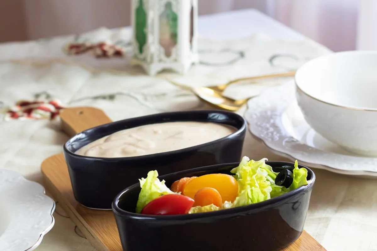Two black wooden bowls on a wooden board with fresh salad greens and a small bowl of dressing on a light table.