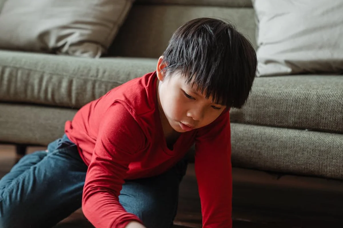 A young child in a red shirt sits on the floor and plays with a wooden toy near a sofa in a living room.