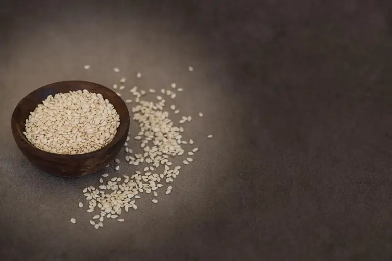 Wooden bowl filled with sesame seeds with seeds spilled on a dark surface