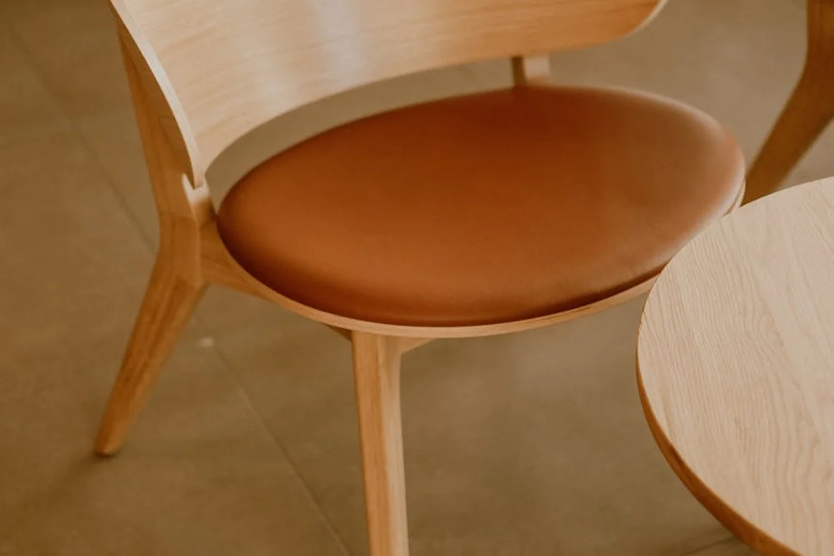 Light wooden chair with a curved backrest and brown leather-like seat beside a round wooden table on a warm-toned floor.