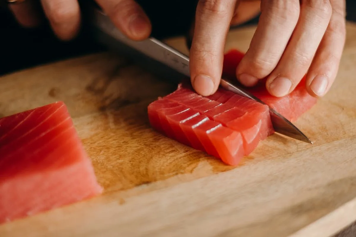 Close-up of a knife slicing a chunk of red tuna on a wooden cutting board.