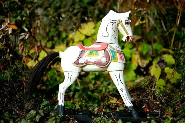 White wooden rocking horse with a colorful saddle standing on grass with green foliage in the background