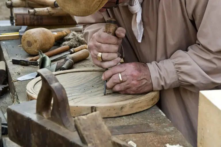 Close-up of a woodworker carving a circular wooden disk on a workbench, surrounded by chisels and woodworking tools.