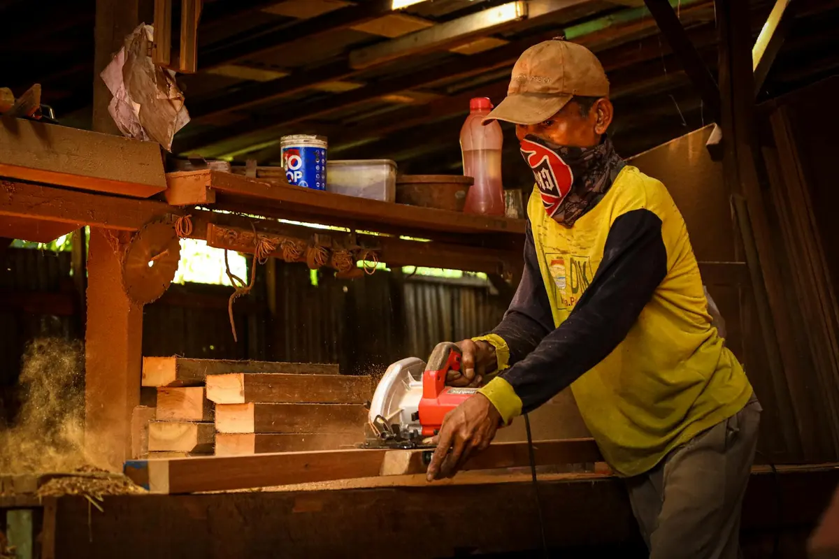 Woodworker wearing a cap and face covering operates a circular saw on a wooden workbench in a dusty workshop.