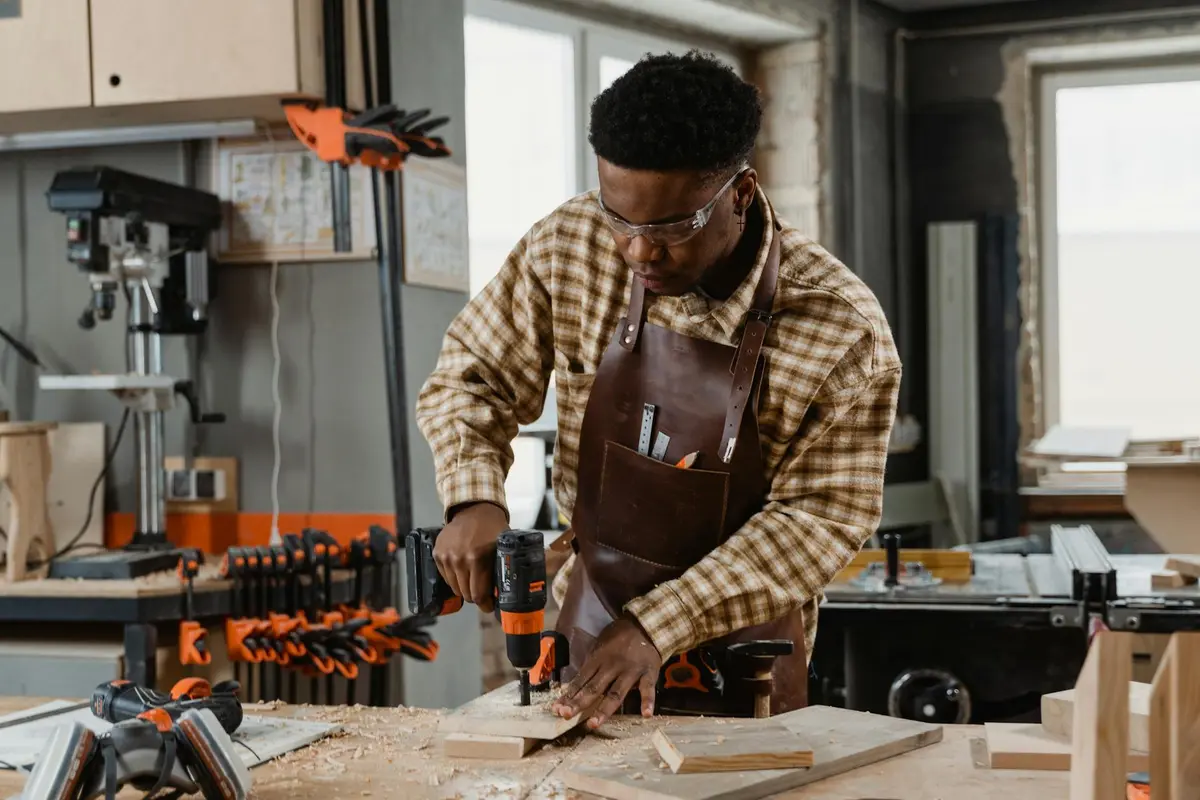 A woodworker wearing safety goggles and a brown apron drills a piece of wood on a workbench, with clamps and tools visible in a workshop.