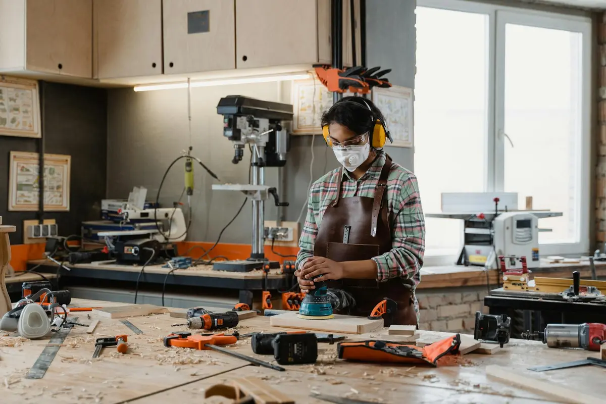 Woodworker wearing a mask and apron stands at a cluttered workbench in a workshop, surrounded by sanding tools.