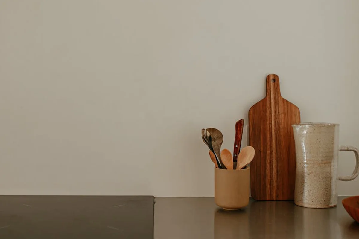 Minimalist kitchen workspace with wooden cutting boards in the background and a cup of wooden utensils on a dark countertop.