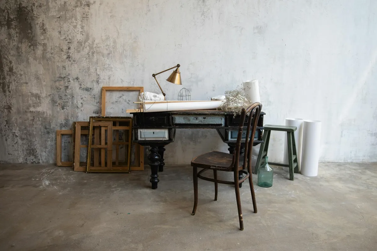 A rustic woodworking studio with a desk and chair, picture frames leaning against a weathered wall, and rolls of paper in the background, highlighting carved and molded wood details.
