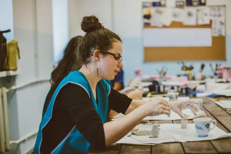 Female craftsworker with glasses and a bun, wearing a teal apron, shaping a ceramic piece at a wooden workbench in a busy studio with tools and cups on the table.